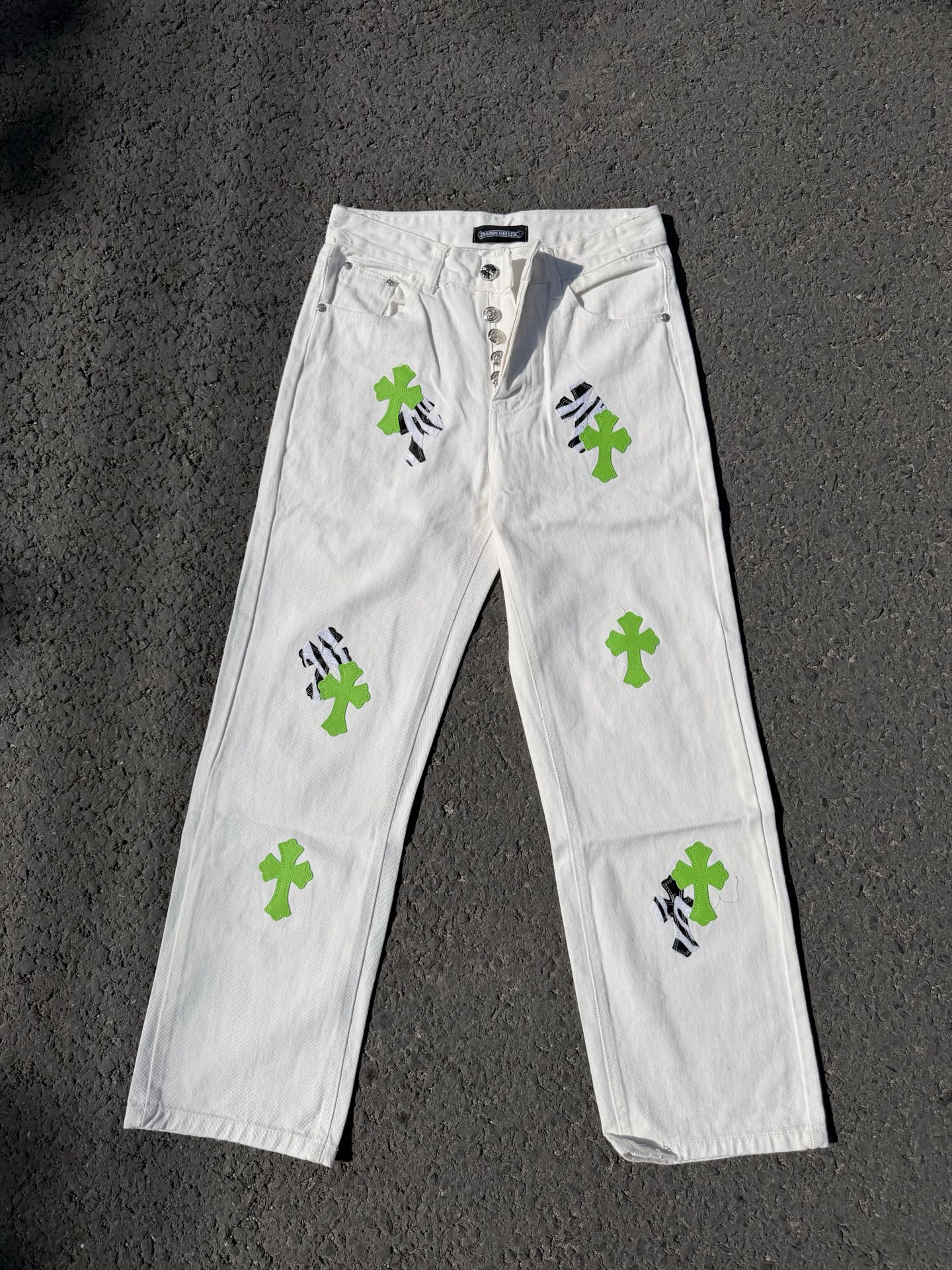 White denim pants with button fly, green cross patches, and black graphic details
