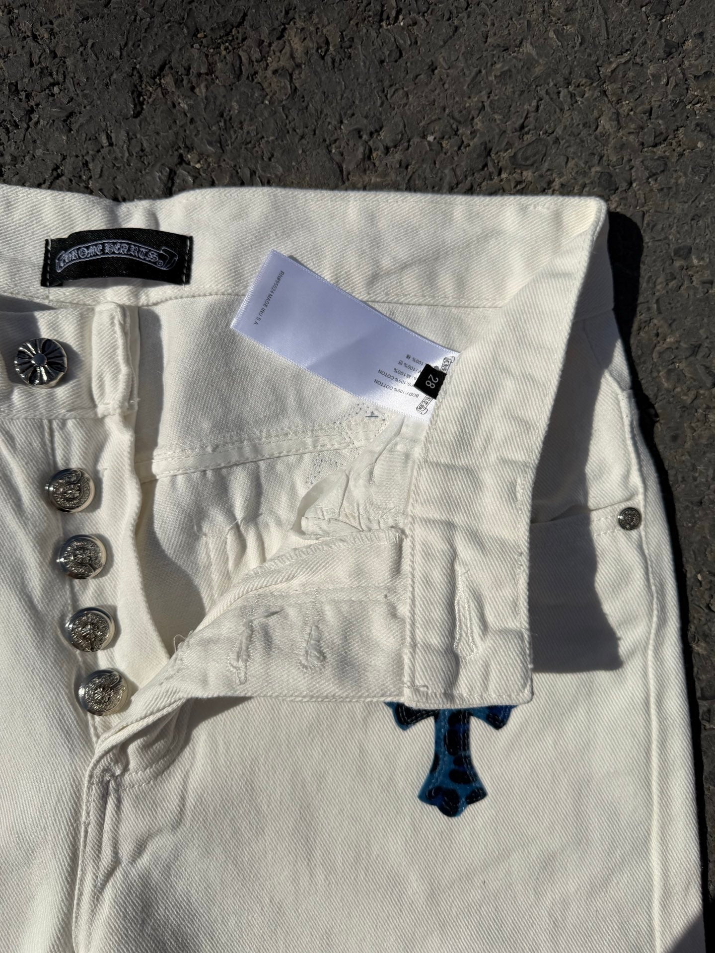 White pants with button fly, front pocket, and orange/blue cross design near hem