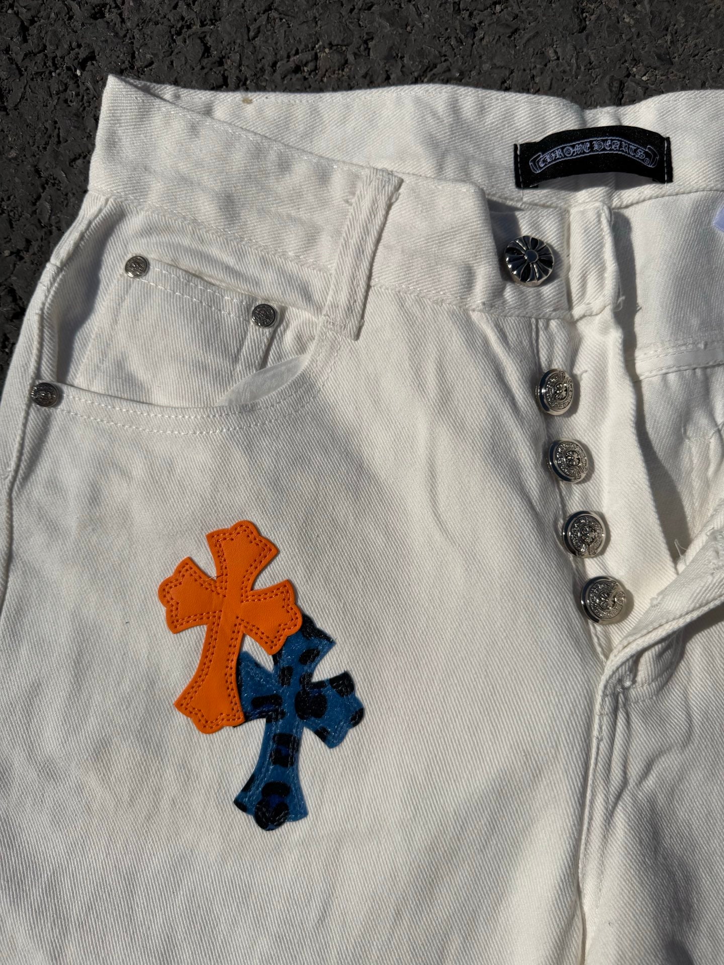 White cross-print pants close-up of button fly, coin buttons, and orange/blue graphic