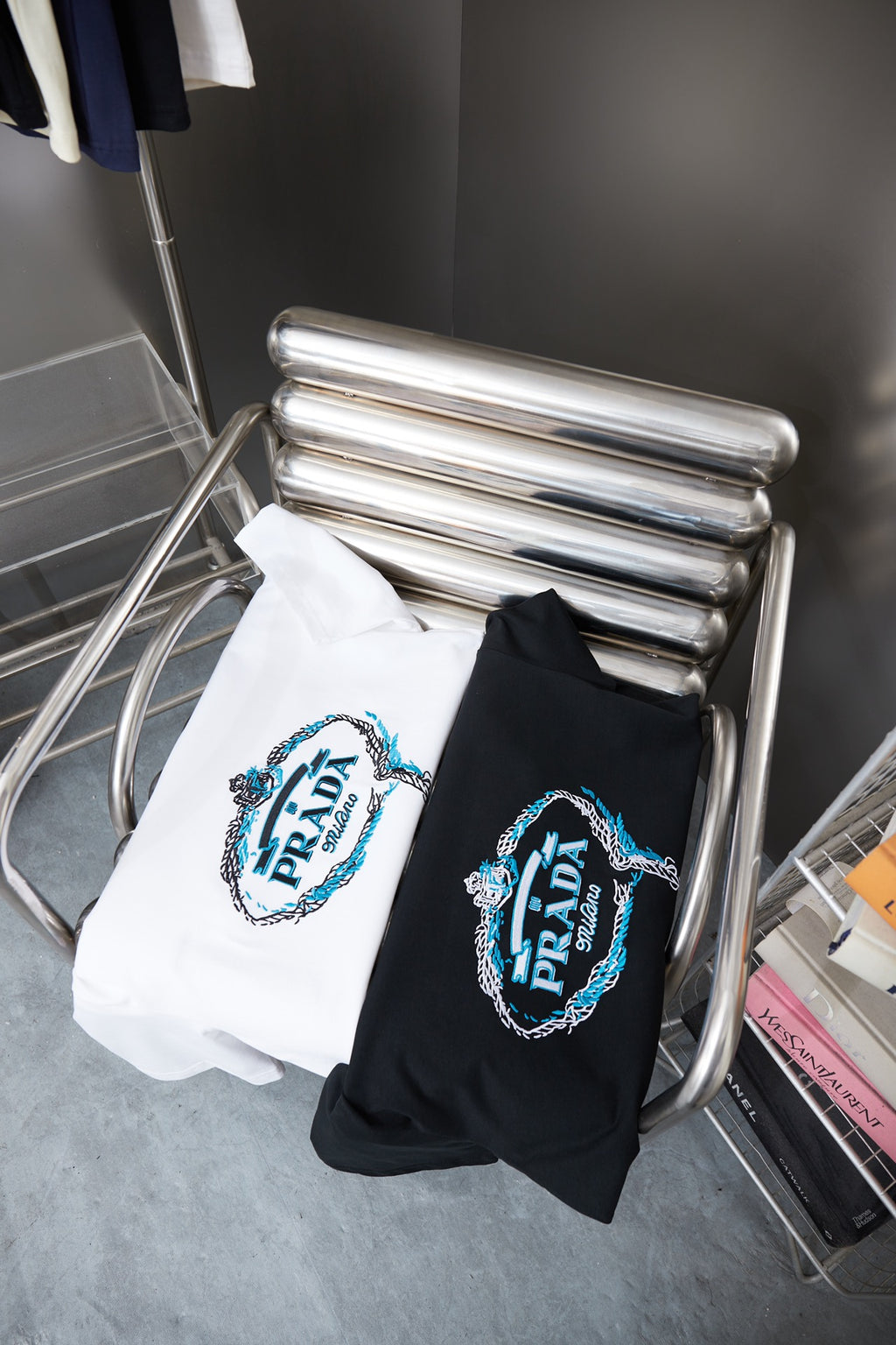 Two folded Prada T-shirts on rack: white and black with embroidered PRADA logos