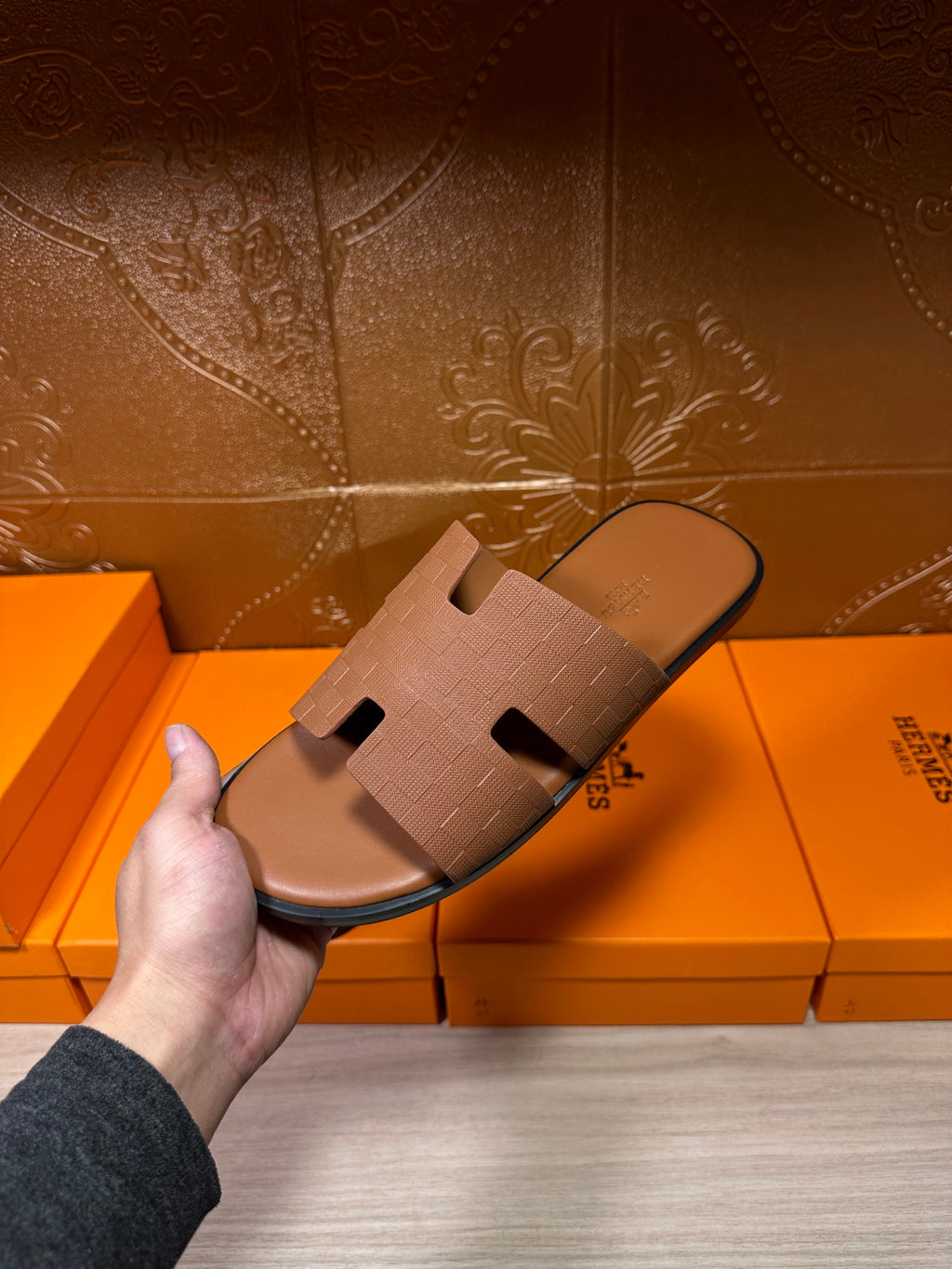 Hermes tan slide sandal with embossed strap and cutout detail, held above boxes