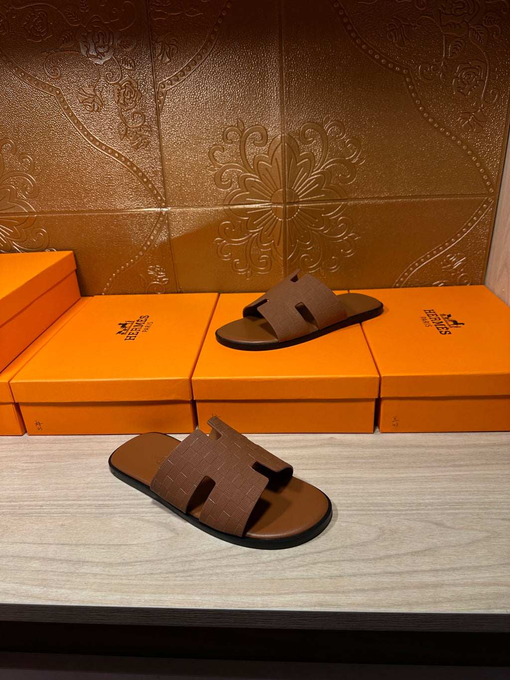 Hermes tan slide sandals with embossed strap, pair shown on display surface near boxes