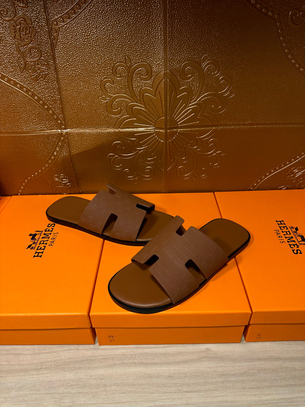 Hermes tan slide sandals with embossed strap, shown on top of orange retail boxes