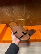 Hermes slide sandal in tan with embossed strap, held in hand above product boxes
