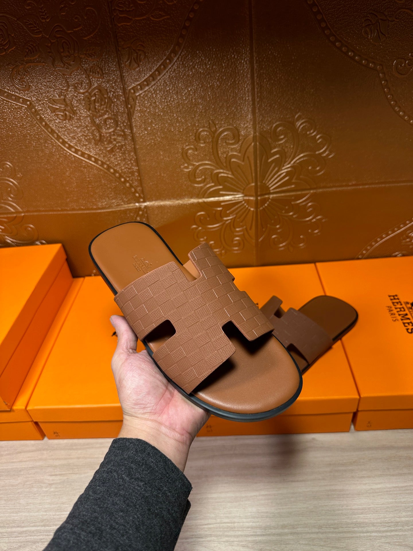 Hermes slide sandal in tan with embossed strap, held in hand above product boxes