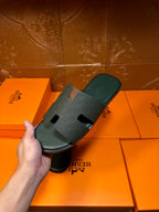Hermes slide sandals held in hand, dark green open-toe slides with H-style strap