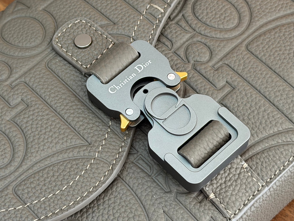 Dior men's bag charm close-up with engraved logo and gold-tone hardware on grey material