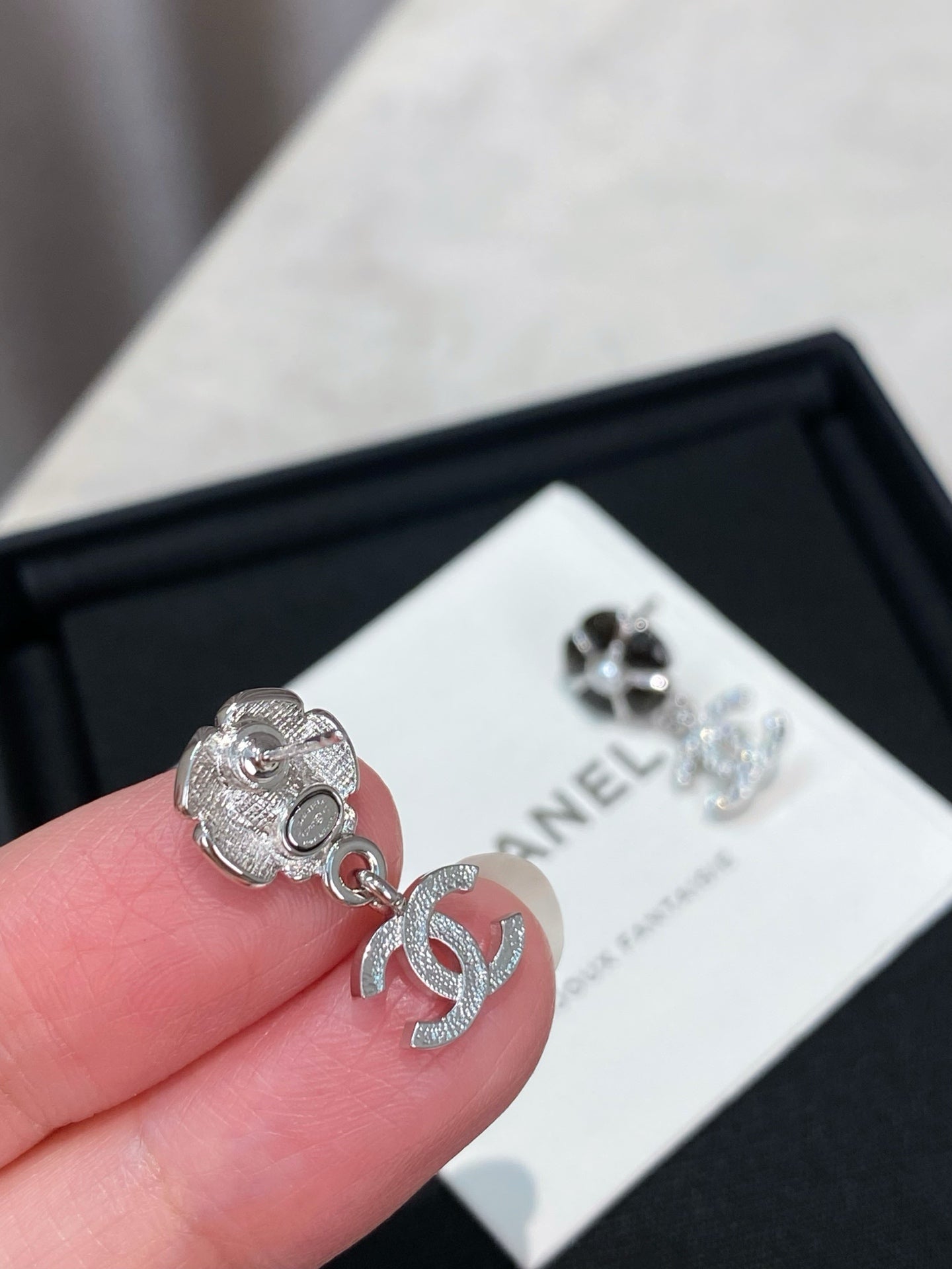 Silver-tone flower stud earring with dangling logo, held near a CHANEL card.