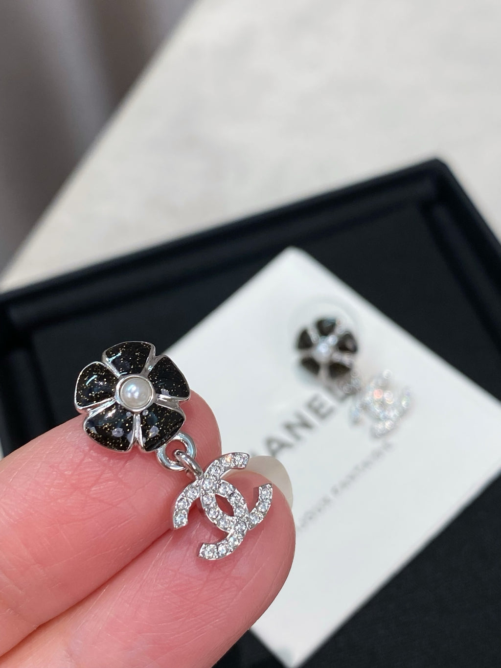 Silver-tone flower stud earring with dangling logo, held near a white CHANEL card.