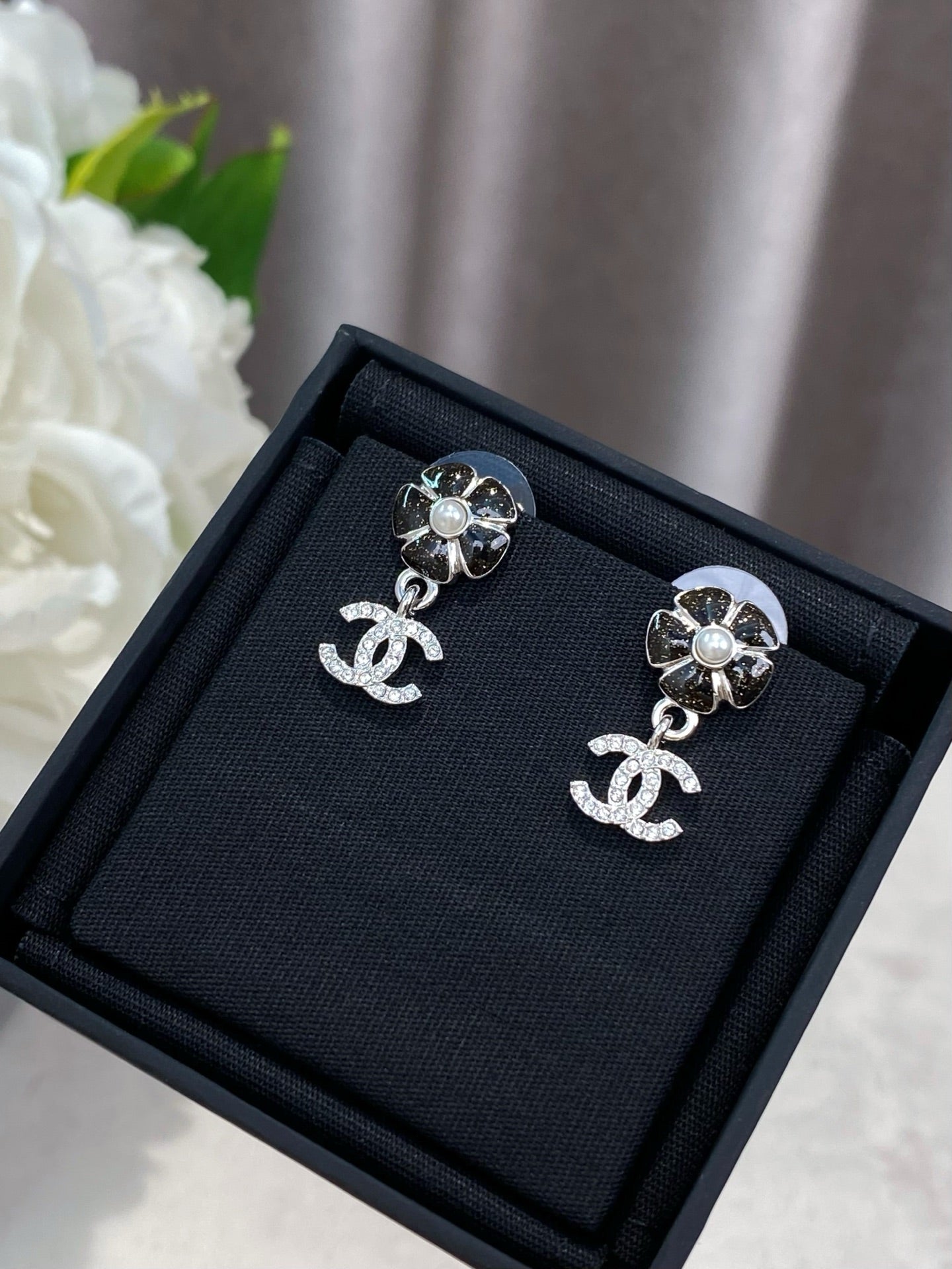 Silver-tone flower stud earrings with dangling logo, laid on black jewelry box.