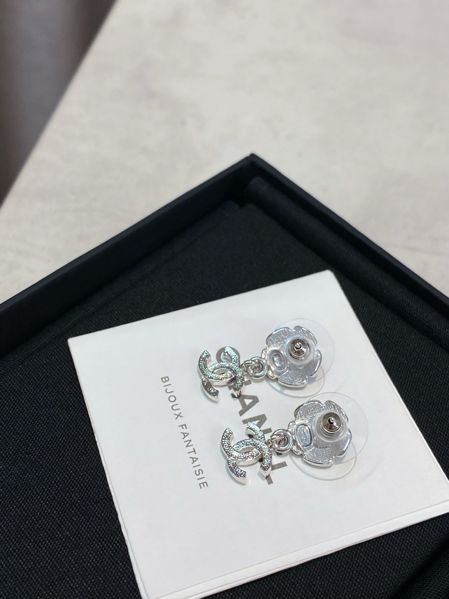 Silver-tone flower stud earrings with dangling logo, on white label inside black box.