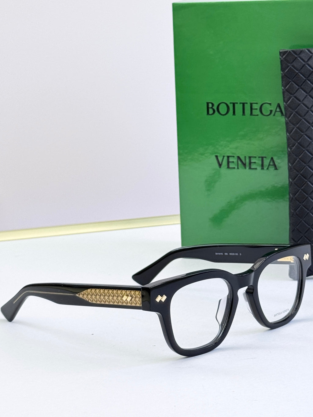 Bottega Veneta eyeglasses with black frames and gold textured side arms, profile view.