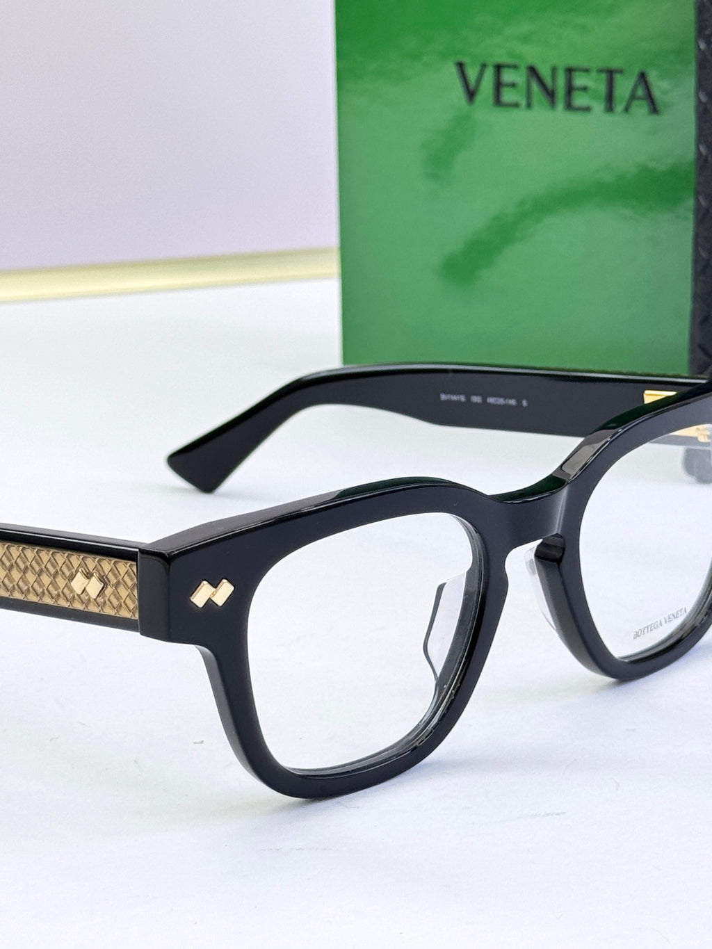 Bottega Veneta eyeglasses with black frames and gold patterned temples, close-up.