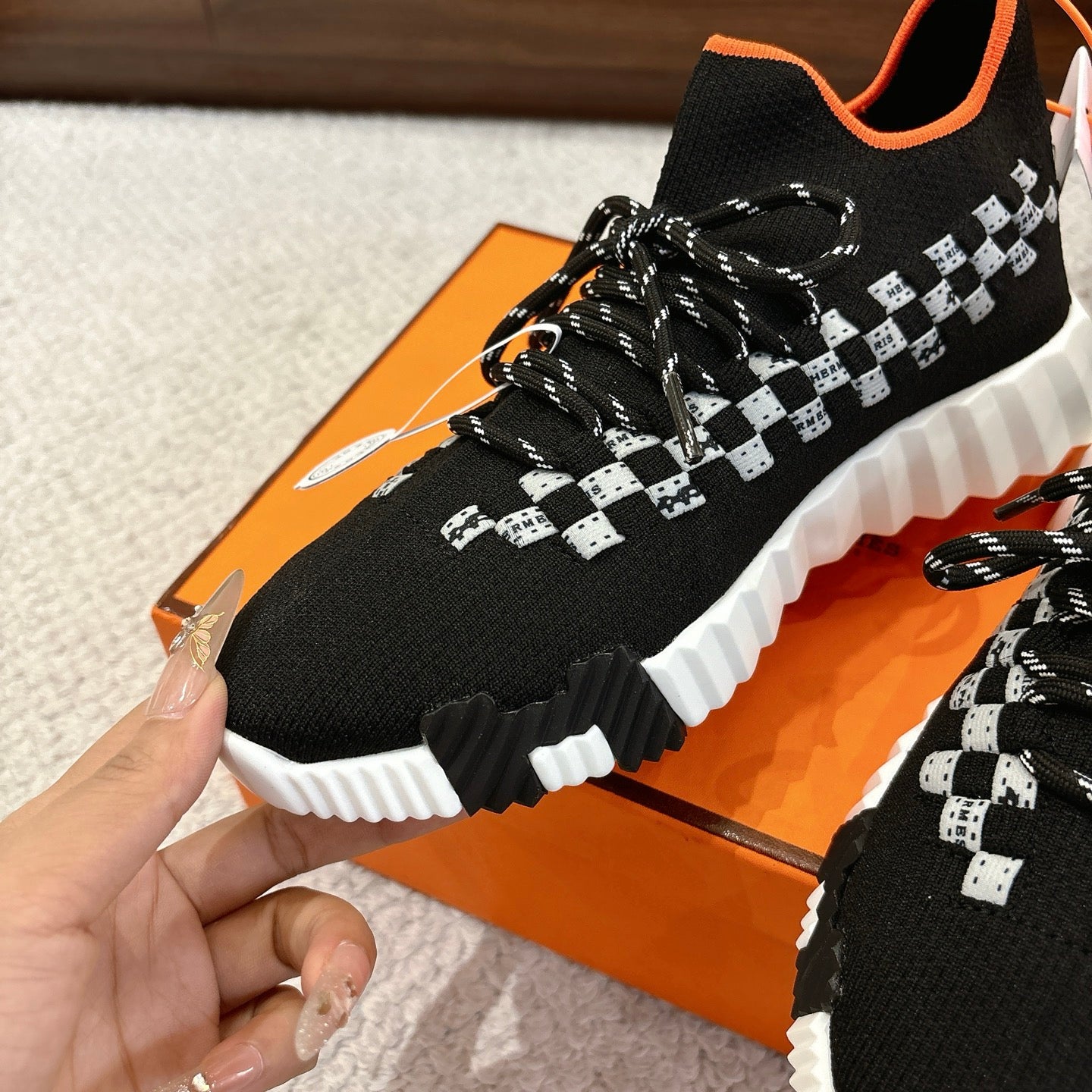 Hermes unisex sneaker close-up: black knit, white checker pattern, orange collar trim, laces