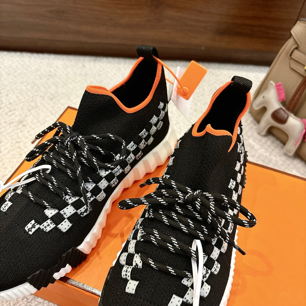 Hermes unisex sneakers close-up: black knit with white check panel, orange trim, braided laces
