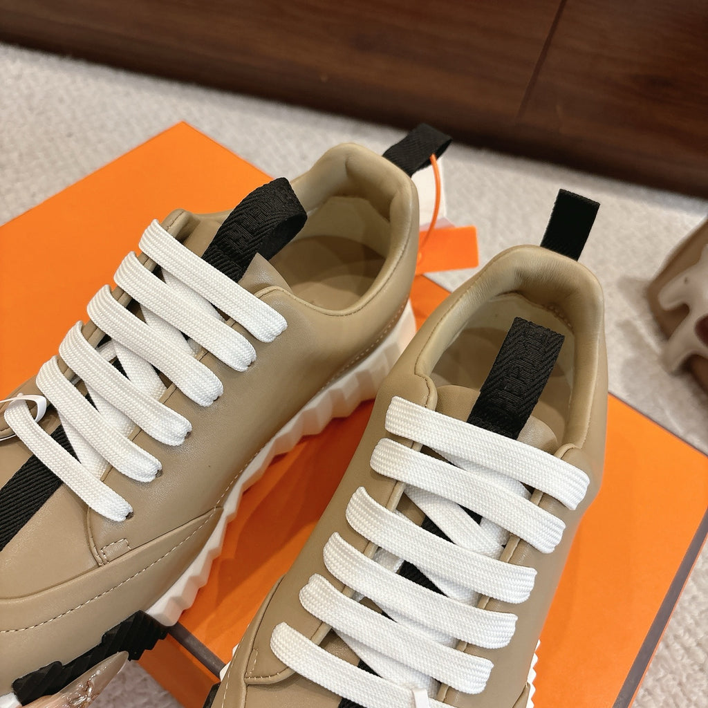 Beige unisex sneakers with white laces, black strap detail, and chunky white tread sole.