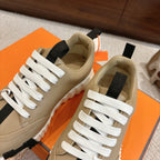 Beige unisex sneakers with white laces, black strap detail, and chunky white tread sole.