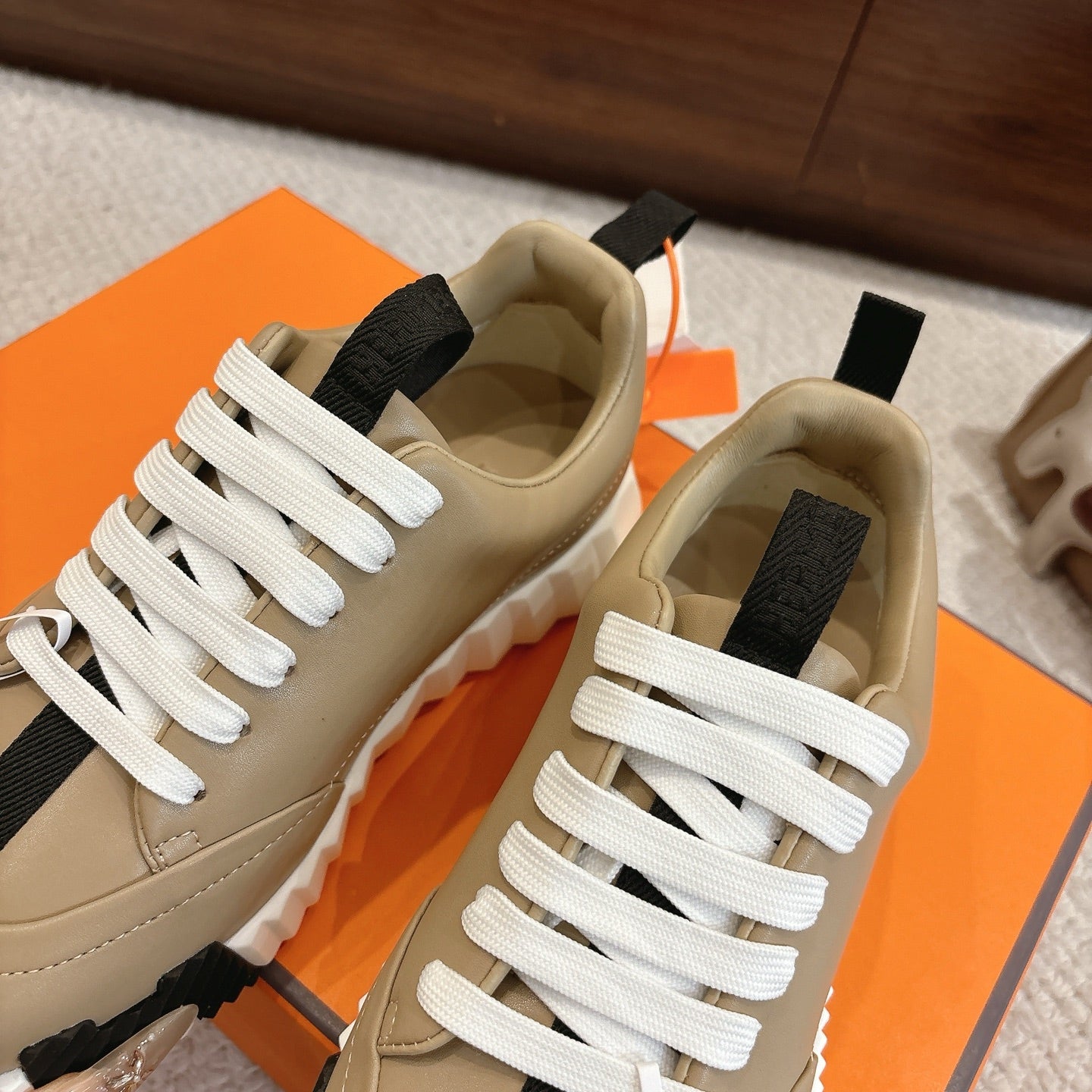 Beige unisex sneakers with white laces, black strap detail, and chunky white tread sole.