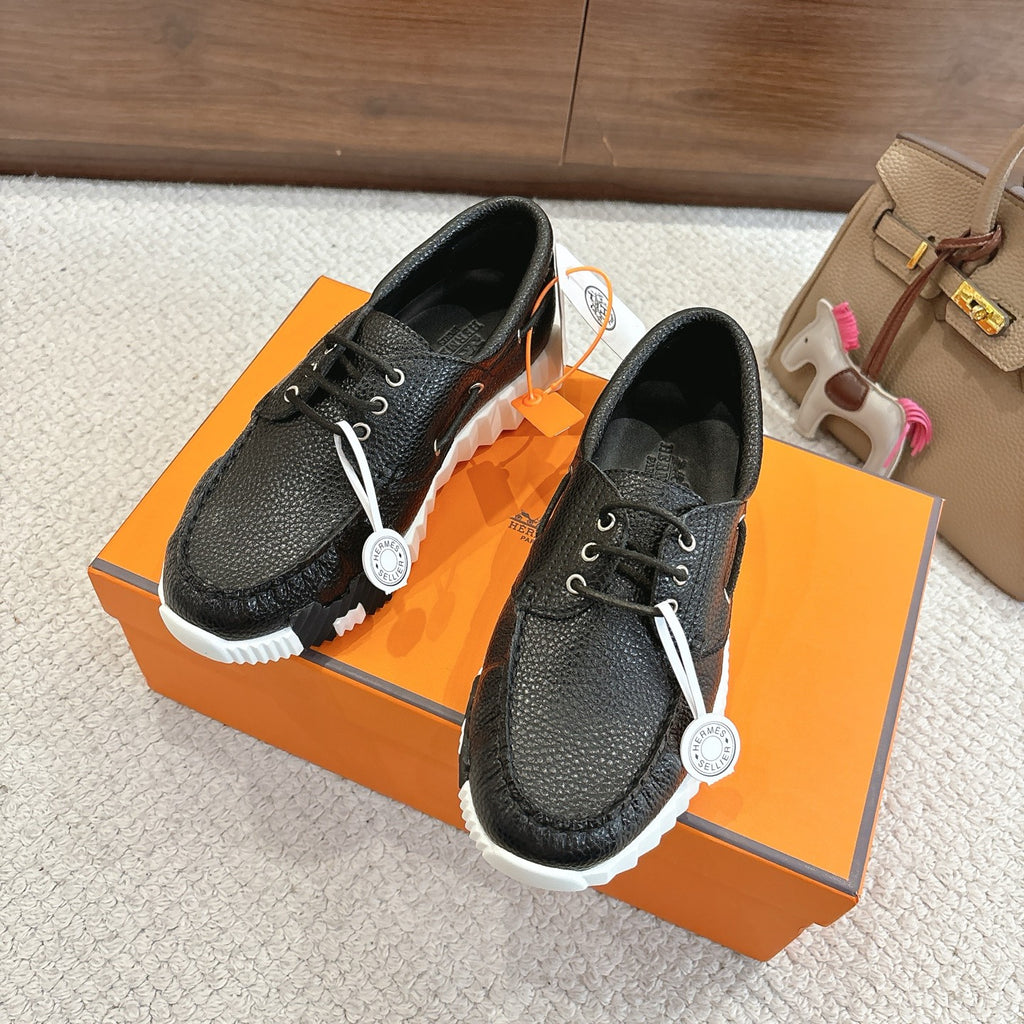 Black leather lace-up shoes with white sole, front and side angle, pair on orange box.