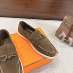 Close-up of dark olive suede loafers with braided trim, metal bit hardware, white sole.