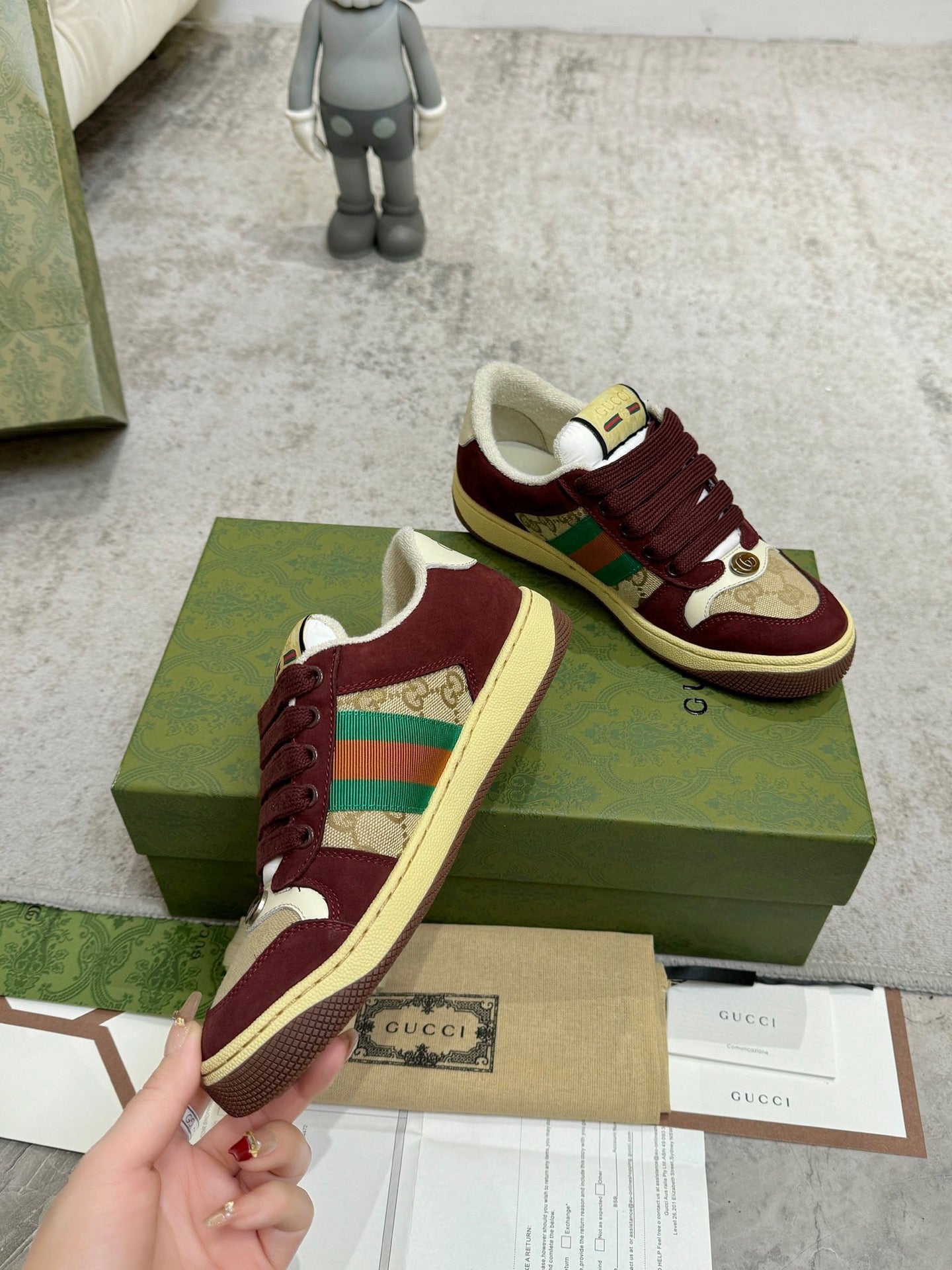 Burgundy Gucci low-top sneakers with striped side panel next to retail packaging