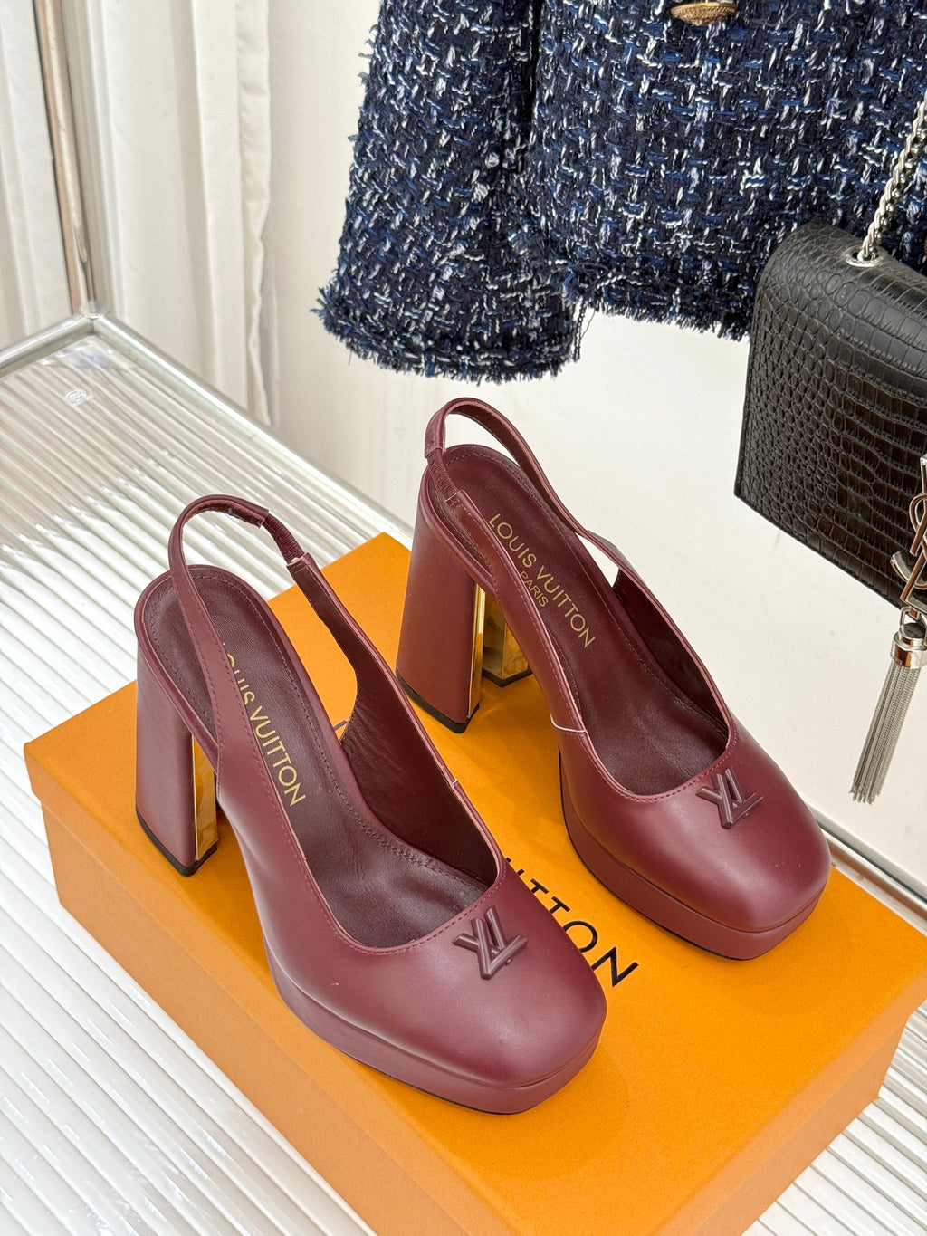 Burgundy leather high-heel pumps with monogram detail, shown on an orange box