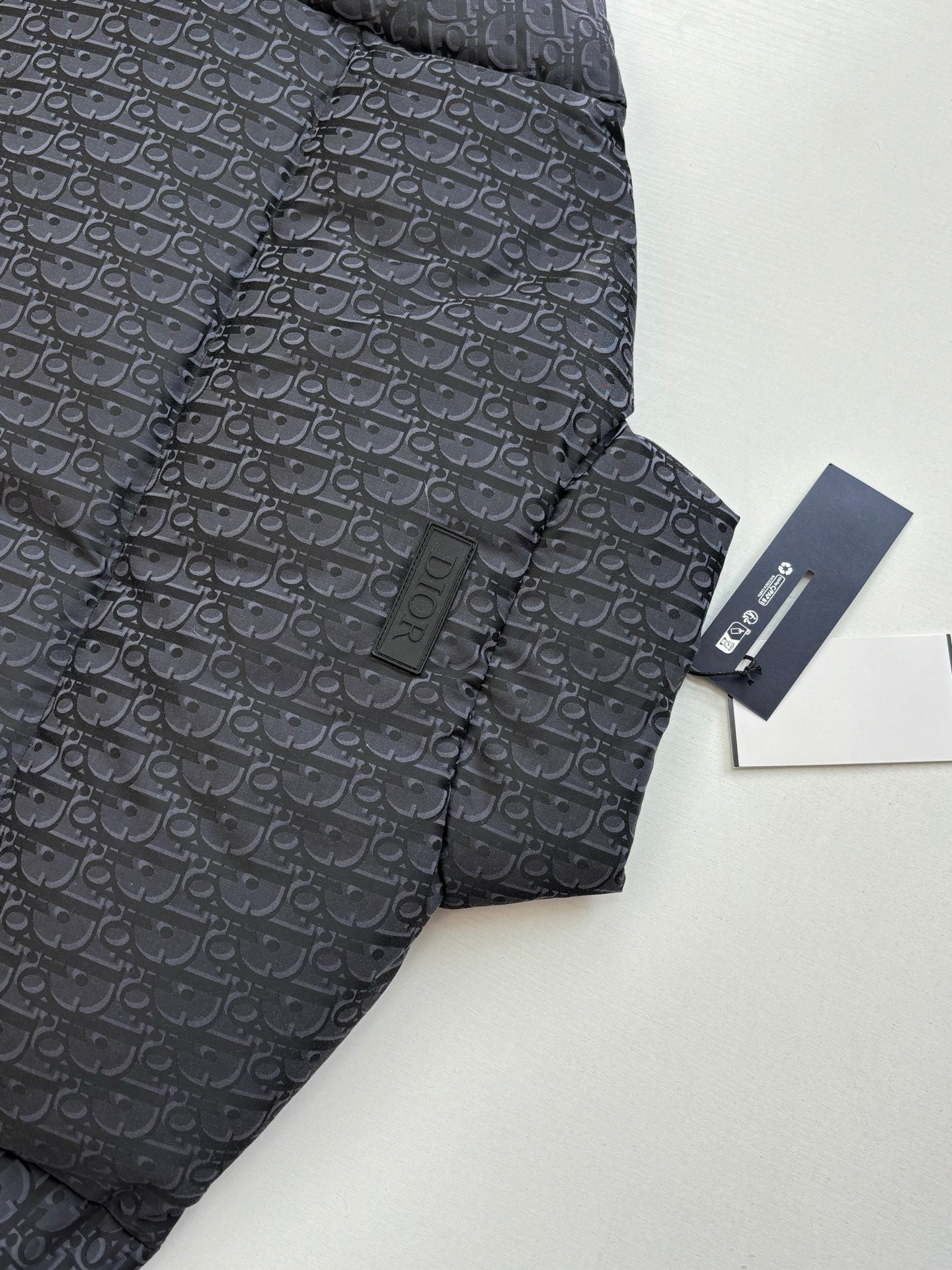 Long quilted Dior jacket with standing collar, dark monogram pattern, tag attached.