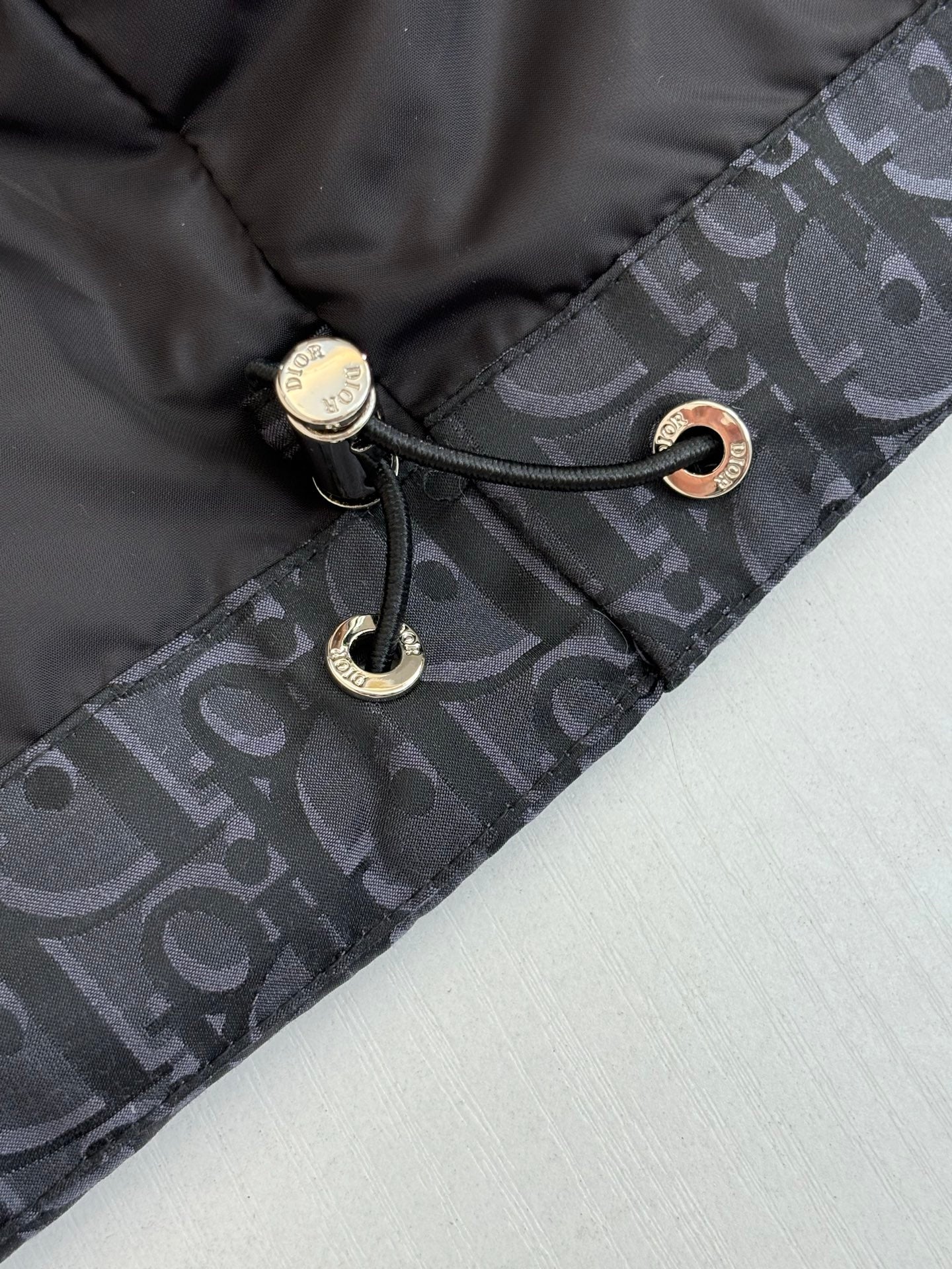 Dior jacket hood drawstring with metal toggles and black cord on dark monogram fabric.