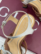 Gucci heeled sandal close-up, metallic silver ankle strap and jeweled buckle, tan footbed