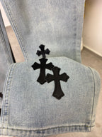 Light wash denim pant leg with large black cross patch at hem