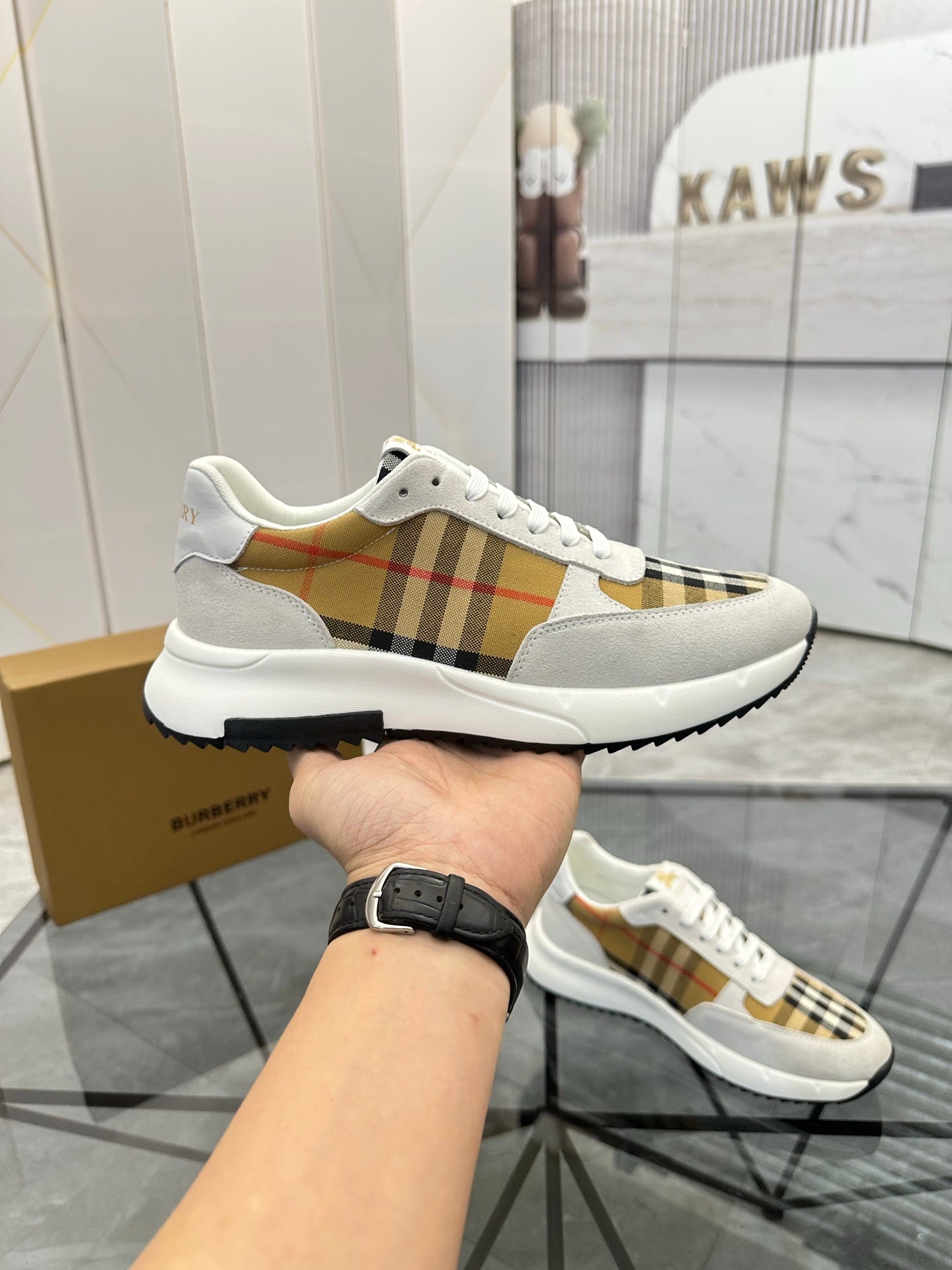 White lace-up sneaker held in hand, tan plaid stripe panel, black outsole tread