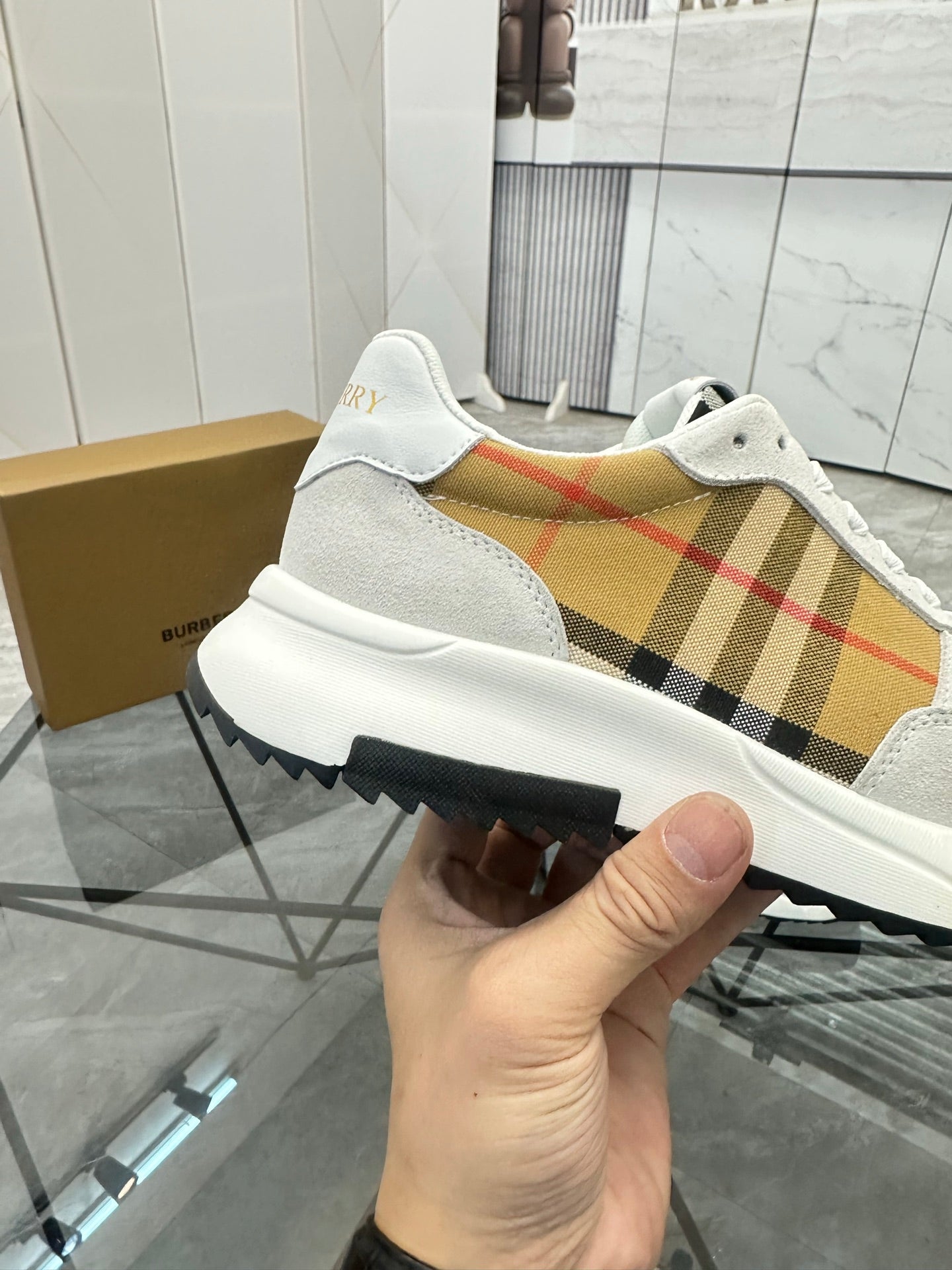 White slip-on sneaker style with tan plaid upper panel, black outsole, held by hand