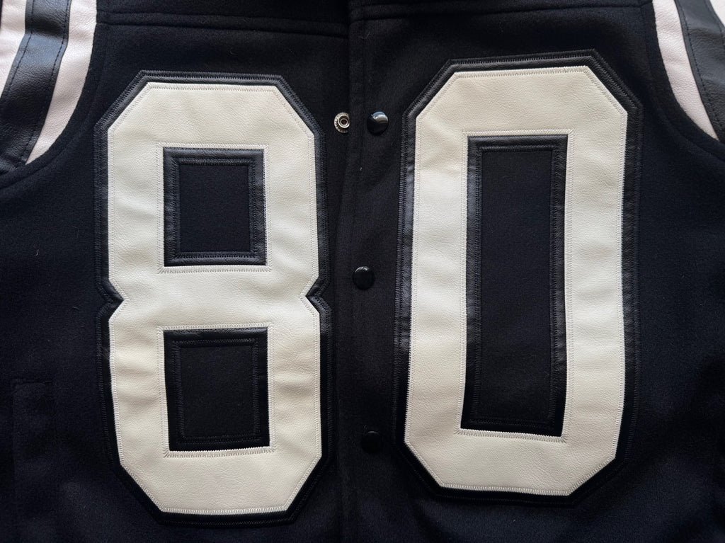 Black varsity jacket front close-up with appliqué “80” panels in white and dark