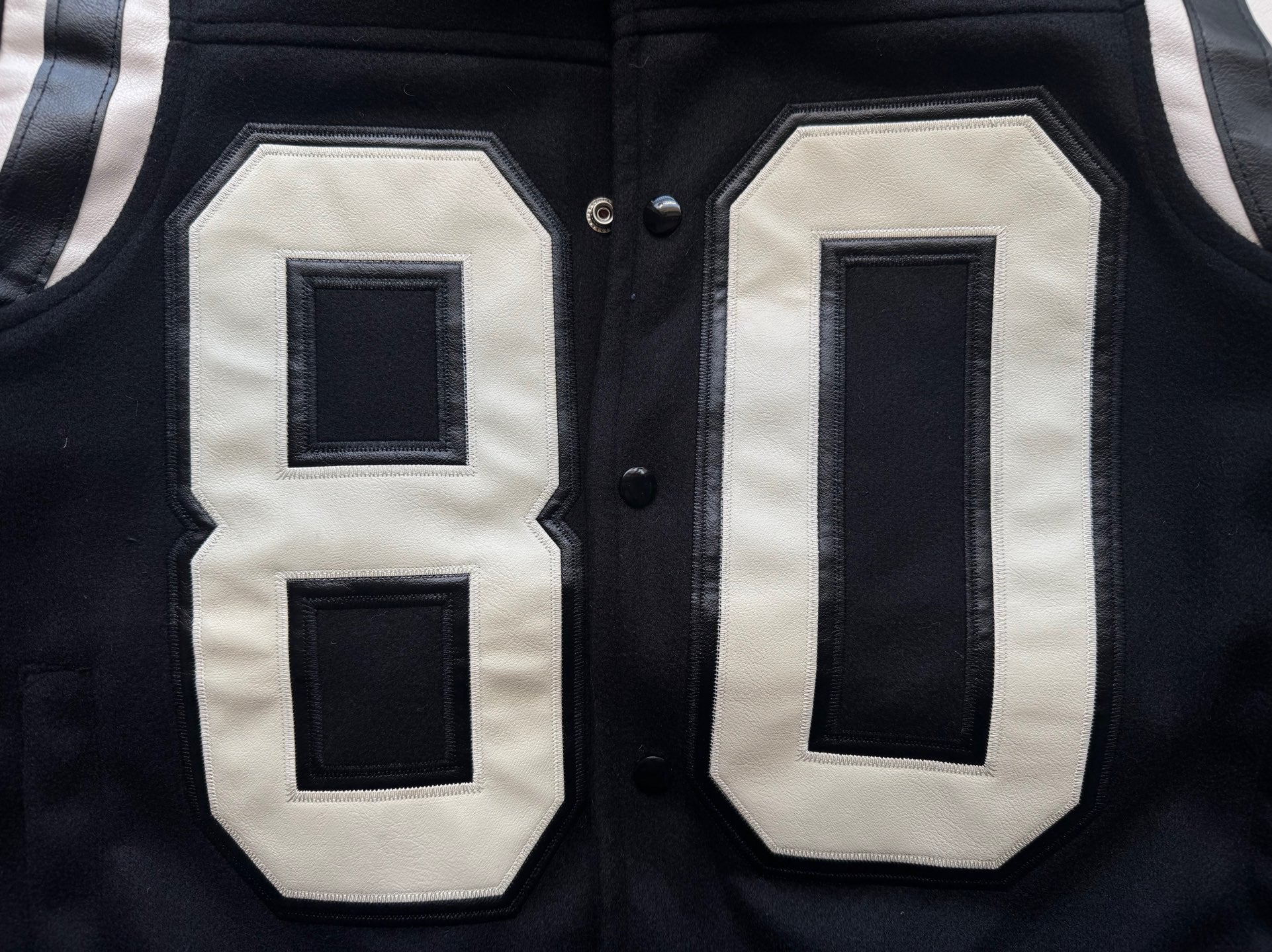 Black varsity jacket front close-up with appliqué “80” panels in white and dark