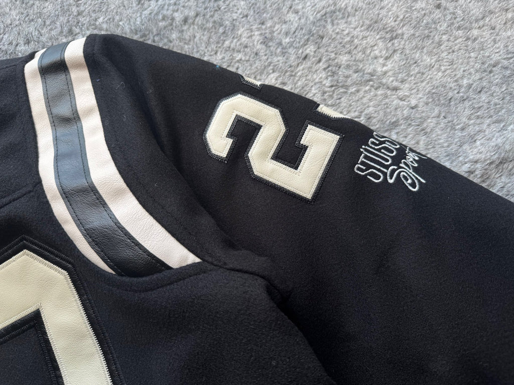 Black varsity jacket sleeve close-up with white stripe, “11” patch, and Stussy script