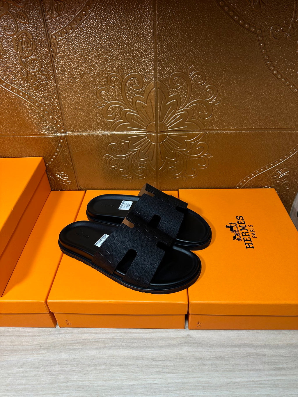 Black slide sandals with textured strap and open toe, laid on orange Hermes box