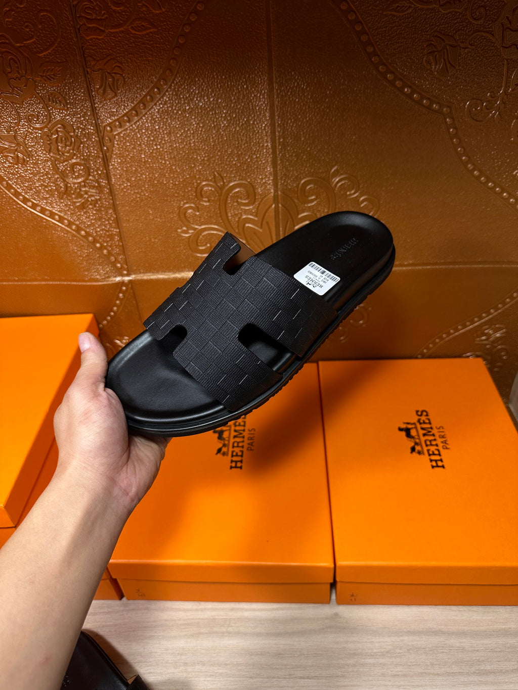 Black slide sandal held in hand above orange Hermes boxes, textured cross-strap