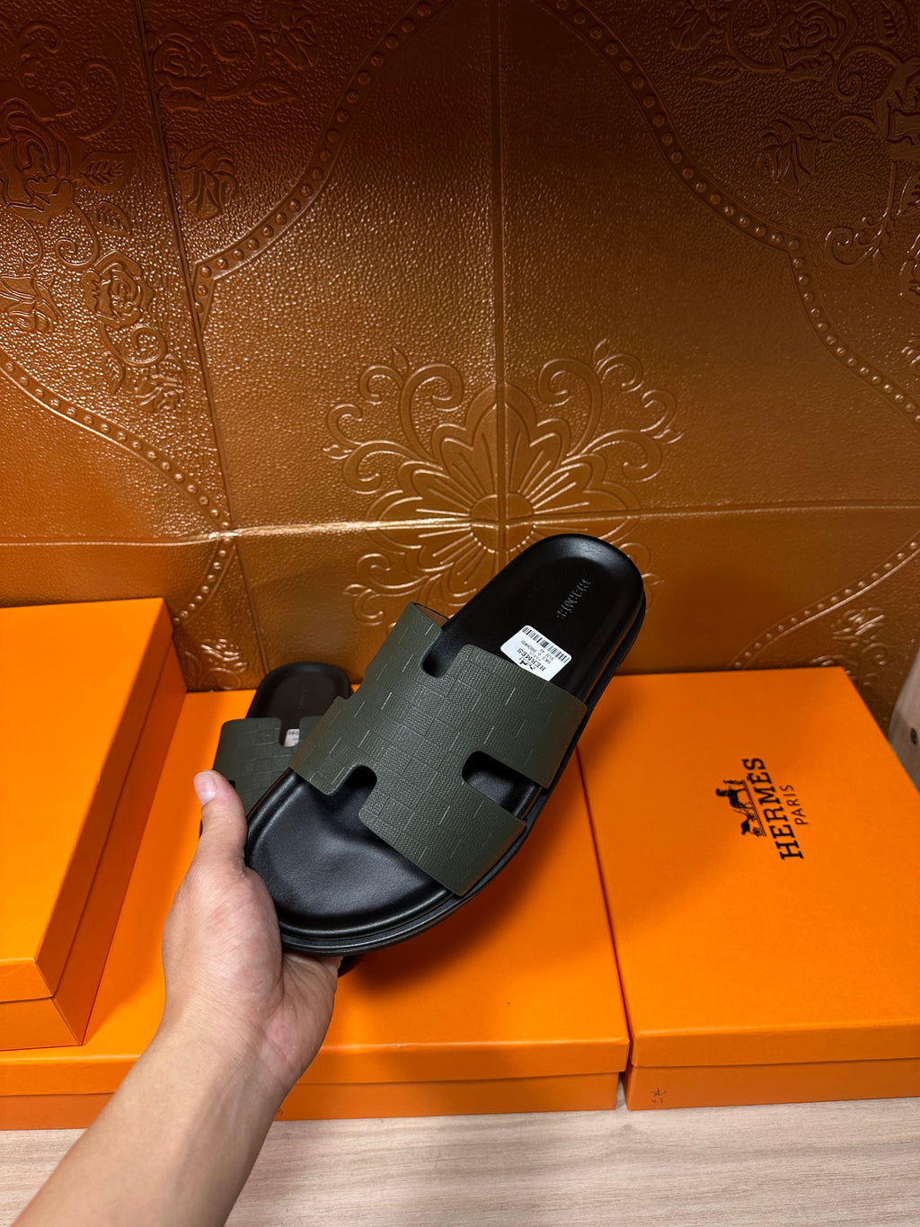 Hermes black slide sandal held beside boxes, textured strap and flat sole