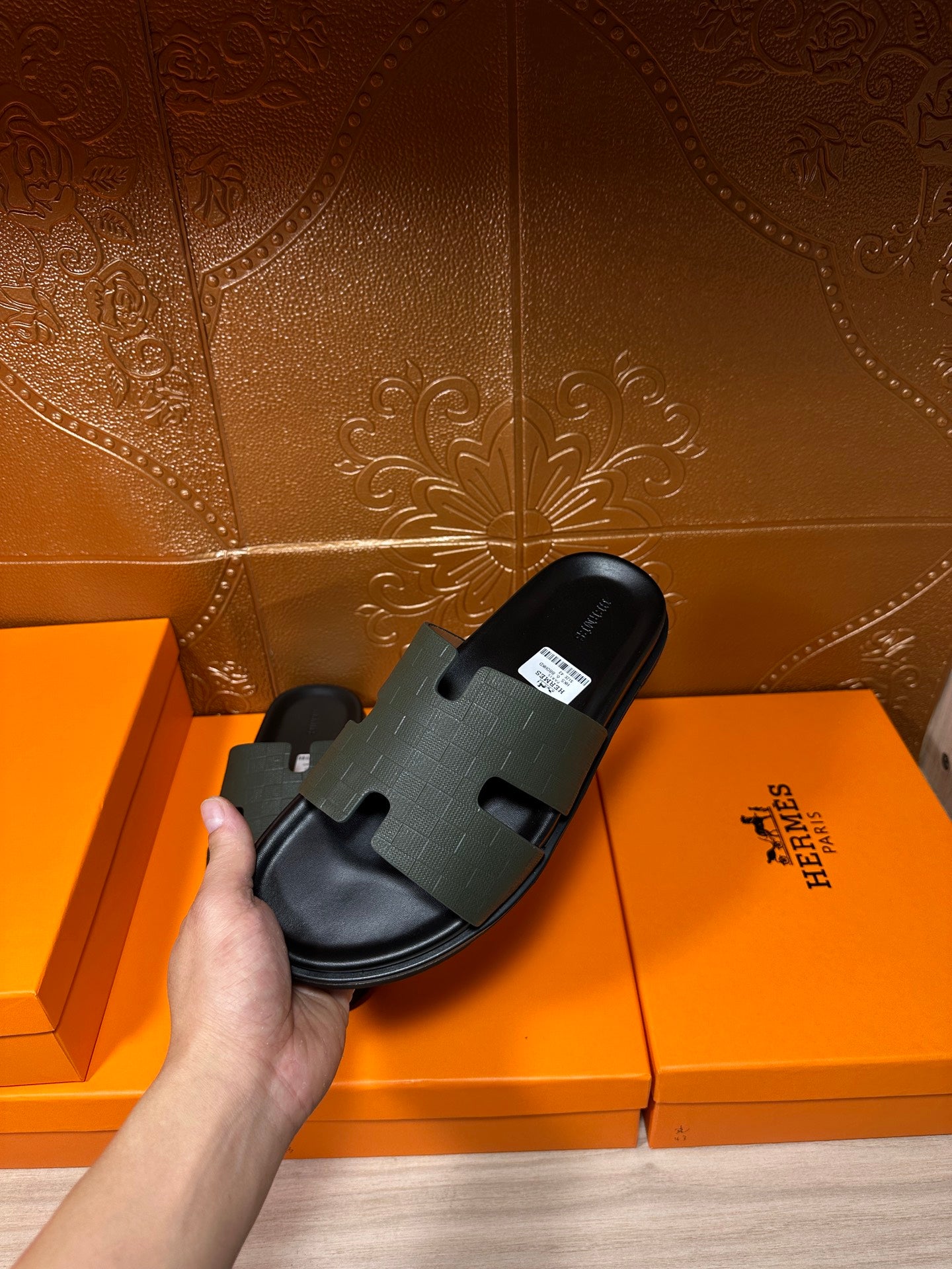 Hermes black slide sandal held beside boxes, textured strap and flat sole