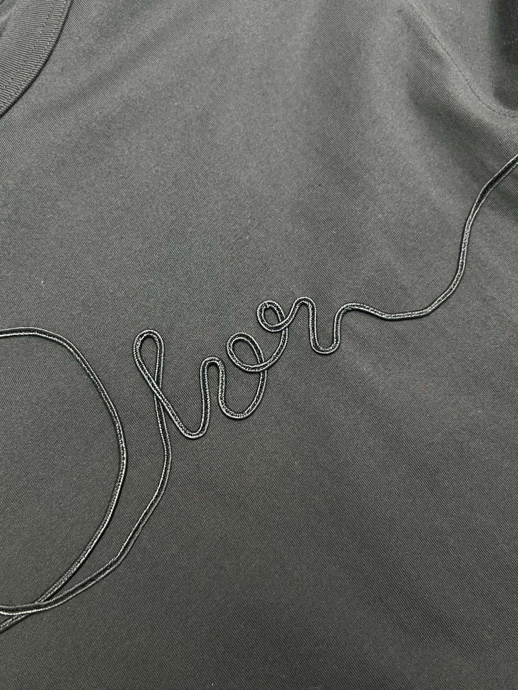 Dior T-shirt embroidery close-up, dark; outlined Dior script near chest