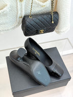 Black heeled pumps with glossy patent toe and Chanel logo on heel, with bag nearby