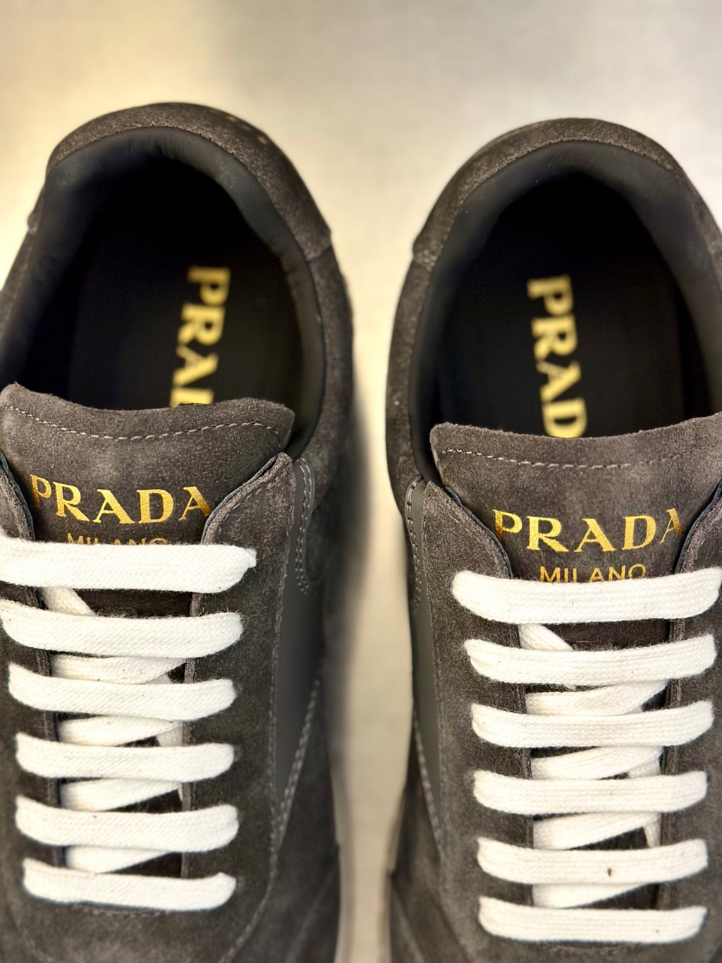 Prada unisex sneakers close-up: dark gray suede, white laces, gold “PRADA” accents