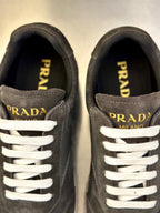 Prada unisex sneakers close-up: dark gray suede, white laces, gold “PRADA” accents