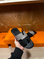 Hermes slide sandal held up: black footbed with patterned gray strap and wide buckle detail