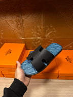 Hermes sandals held in hand over orange boxes; black straps and blue patterned footbed.