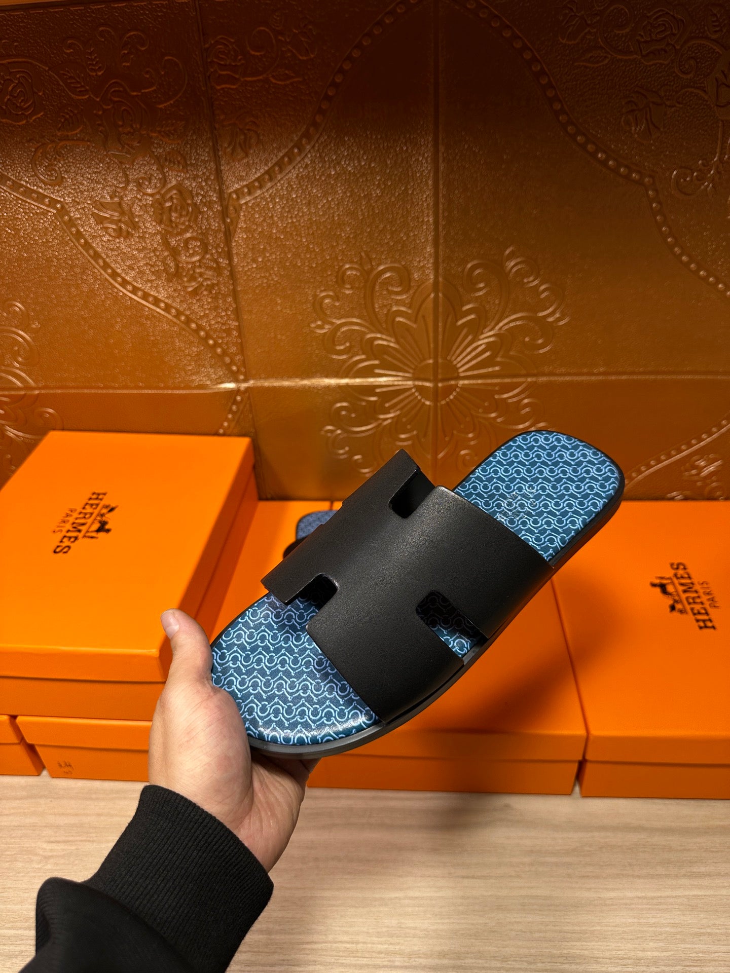 Hermes sandals held in hand over orange boxes; black straps and blue patterned footbed.