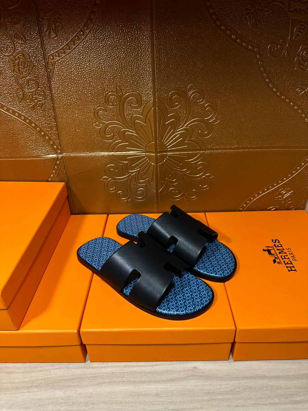 Hermes sandals, black slide straps over blue patterned footbeds, on orange storage boxes.