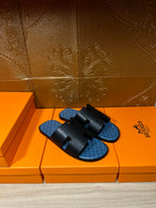 Hermes sandals, black slide straps over blue patterned footbeds, on orange storage boxes.