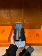 Hermes sandals held from the side: black slide straps and blue patterned footbed.