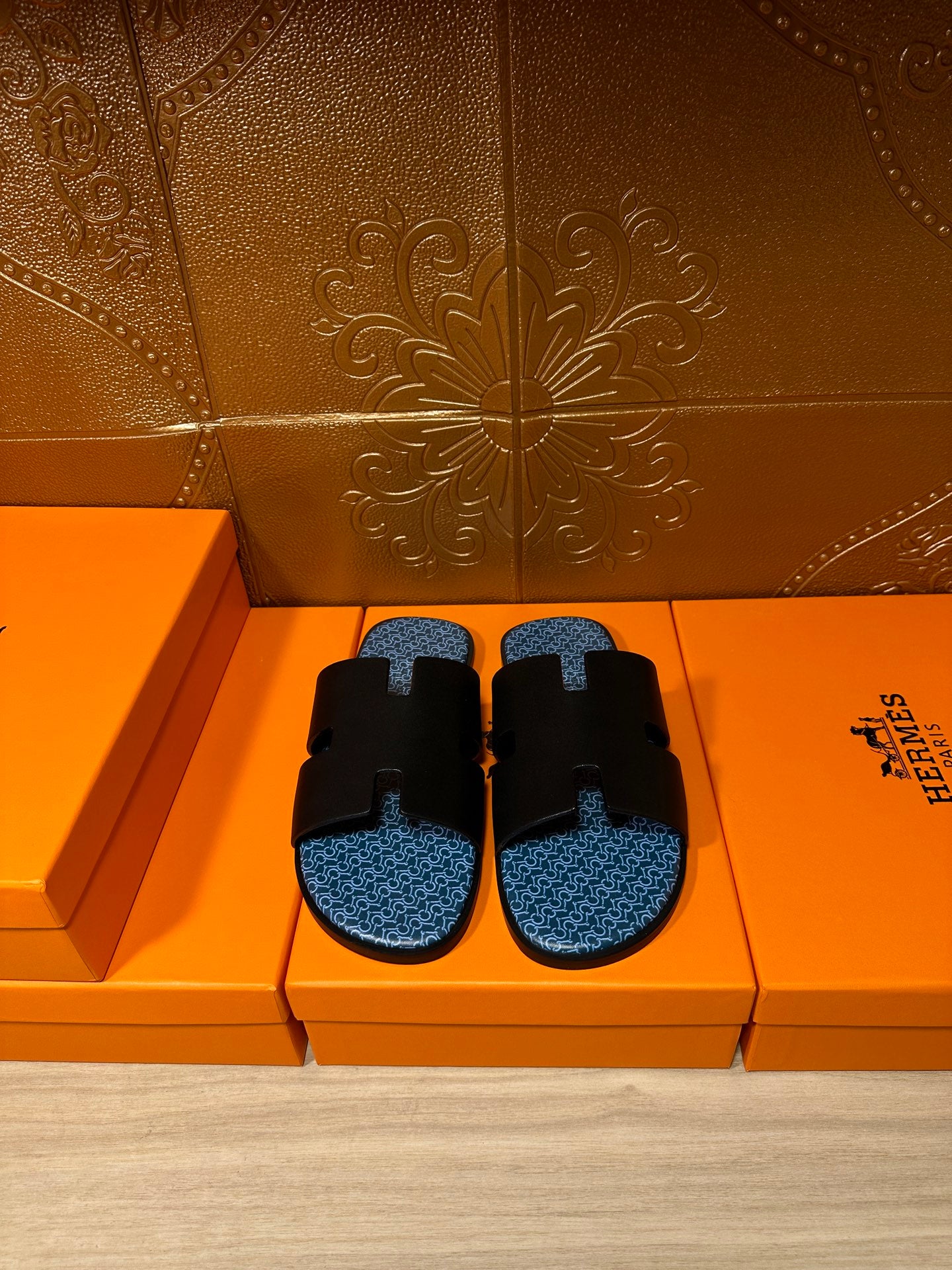 Hermes sandals with black straps and blue patterned footbeds, shown on a box display.